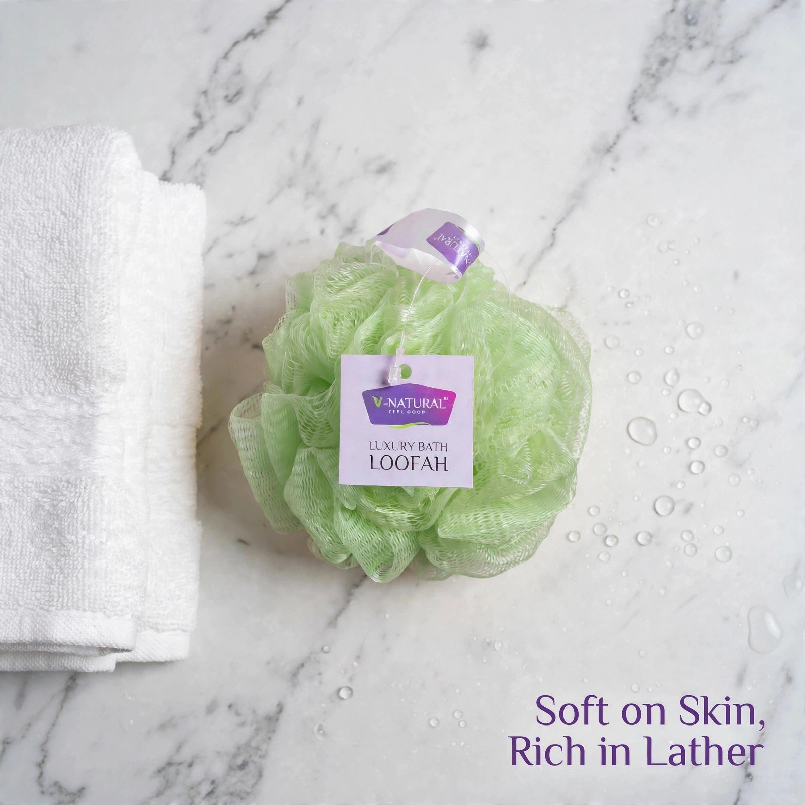 A light green V Natural loofah bath sponge with a white and purple label sits on a white marble surface next to a folded white towel. Purple text in the bottom right corner reads 'Soft on Skin, Rich in Lather'.